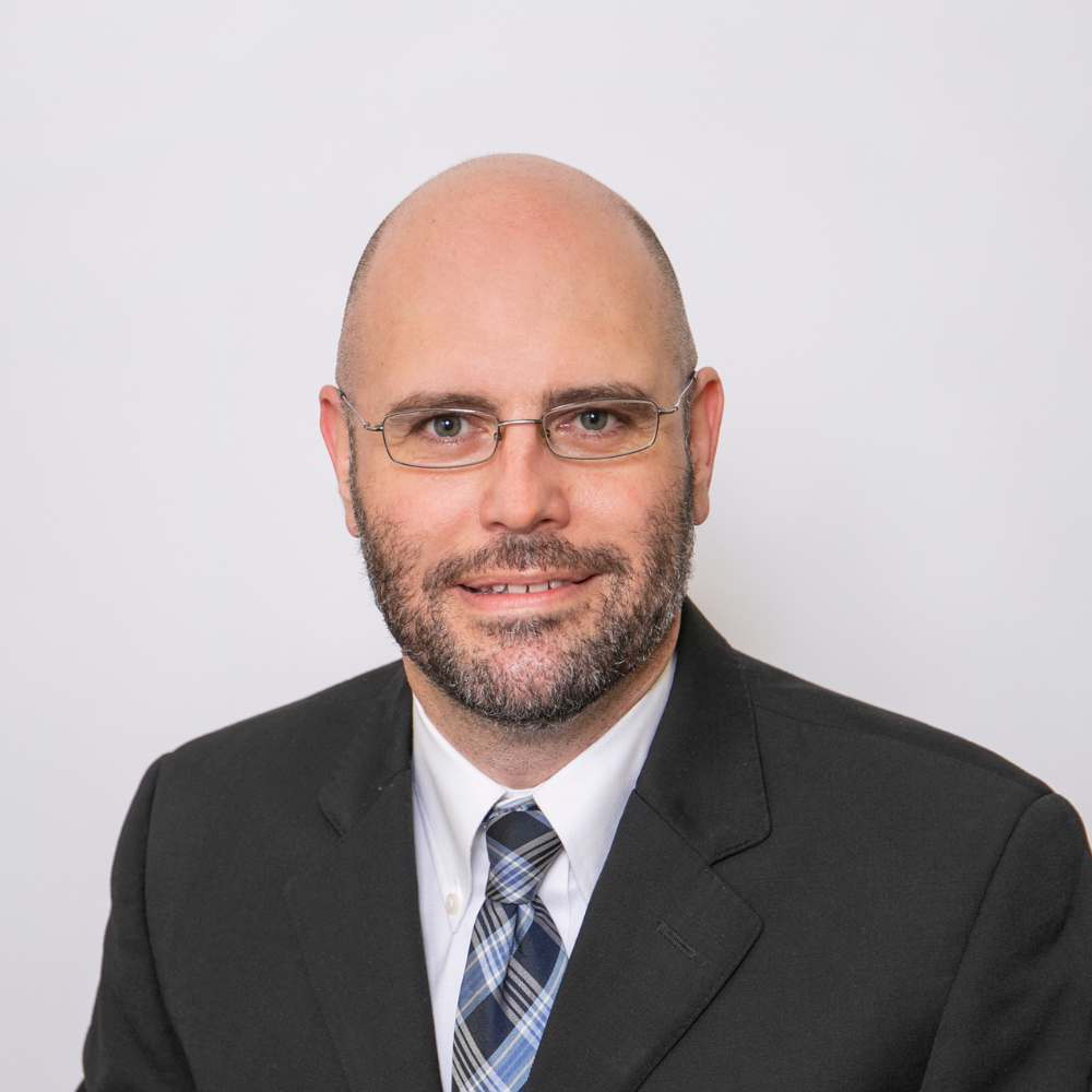 a middle aged, white, bearded man in a grey suit jacket and white collared shirt